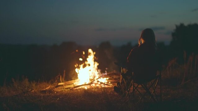 Lonely woman sitting on chair by the fire in the dark. Blonde girl watching fire in nature. Wild camping with bonfire, travel concept