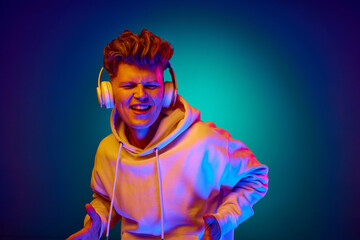 Portrait of funny young guy, man, dressed white hoodie listening music in headphones against gradient mint background in mixed colors neon filter.