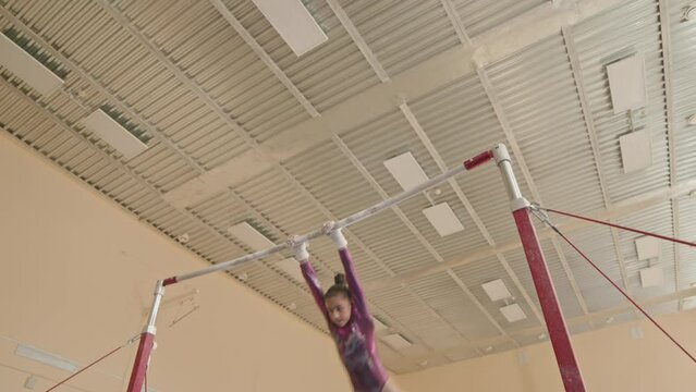 Acrobatic little girl in purple leotard practicing artistic elements on uneven bars in spacious gymnastics studio