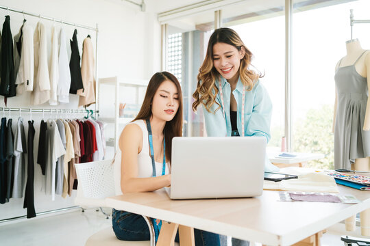 Two Asian female fashion designers or dressmakers brainstormed and discuss the design of the new collection with examples of fabrics and sketches on the laptop. Working together in the studio.