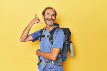 Middle-aged hiker with backpack in studio showing a mobile phone call gesture with fingers.