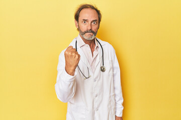 Doctor with stethoscope in yellow studio showing fist to camera, aggressive facial expression.