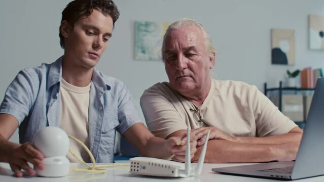 Medium shot of young grandson helping his senior grandfather to install and connect webcam with laptop and Wi-Fi router