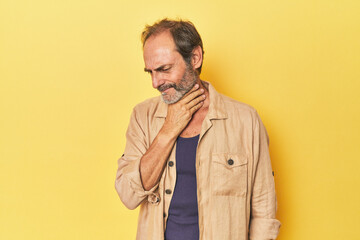 Caucasian middle-aged man in yellow studio suffers pain in throat due a virus or infection.