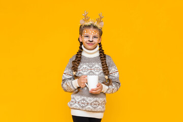 A little girl dressed as a Christmas reindeer holds a cup of hot cocoa in her hands. A child in a sweater is enjoying the holiday. Yellow isolated background.