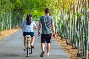 Happy Asian couple enjoy with bicycle in bamboo park. Asian lovers ride bicycle with having fun to exercise activity together in garden. Chilling and relaxing family enjoys nature. Selective focus.