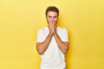 Young Caucasian man on a yellow studio background laughing about something, covering mouth with hands.