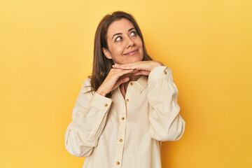 Middle-aged caucasian woman on yellow keeps hands under chin, is looking happily aside.