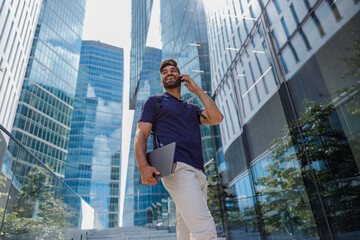 Smiling male entrepreneur standing with laptop and talking phone on skyscrapers background