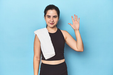 Sporty woman with towel on blue studio smiling cheerful showing number five with fingers.
