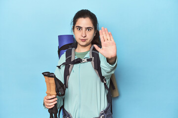 Hiker with backpack and poles on blue standing with outstretched hand showing stop sign, preventing...