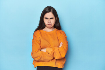 Young Caucasian woman on blue backdrop frowning face in displeasure, keeps arms folded.