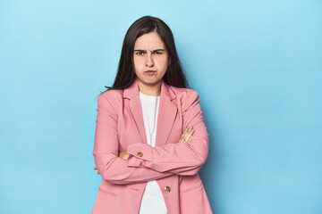 Young Caucasian woman on blue backdrop frowning face in displeasure, keeps arms folded.