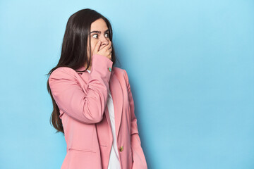 Young Caucasian woman on blue backdrop thoughtful looking to a copy space covering mouth with hand.