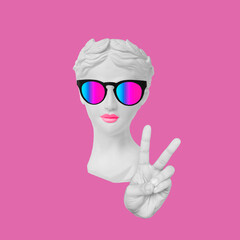 Antique female statue's head in bright sunglasses showing a peace gesture with hand isolated on pop...