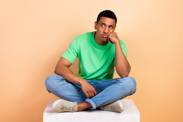 Photo of unhappy disapointed guy dressed green t-shirt sit on platform fist on cheek look empty space isolated on beige color background