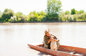 Tender fisherman in hat hugging nice spotted dog, while floating in rowling boat on lake. Front...