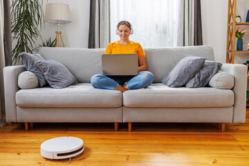 Robotic vacuum cleaner cleaning the living room while woman sitting on sofa