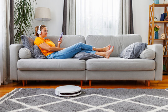 Robotic vacuum cleaner cleaning a room while woman relaxing on the sofa at home