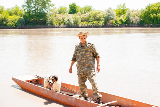 Young caucasian guy standing in shubby boat collecting tackle after fishery outdoors. Front view of male angler floating with dog, with calm, muddy river on background. Concept of fishery.