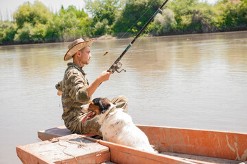 Calm male fisher throwing hook into water, while sitting in rowing boat with dog in hot day. Side...