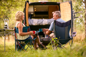 Rear View Of Senior Couple Camping In Countryside With RV Drinking Coffee By Outdoor Fire