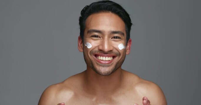 Face, man and apply of moisturiser in studio on gray background for beauty, skincare or glow in mockup. Asian, model and smile for product, sunscreen or protection by hand, hydration or anti aging