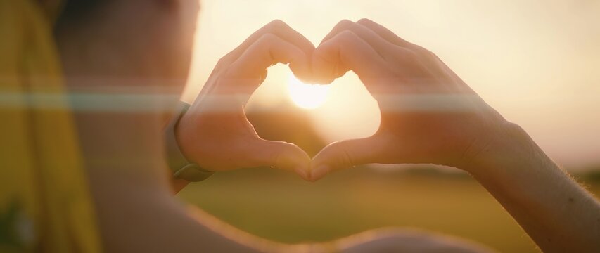 Woman hands showing heart symbol on sunrise golden sky in summer. Close up love, romantic, positive concept.