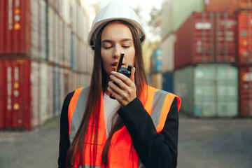 Woman engineer speak to walkie-talkie at shipping containers terminal. Woman in white helmet and vest working - obrazy, fototapety, plakaty