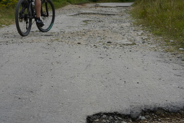 Old asphalt road with many potholes and the cyclist which is trying to avoid the obstacles riding on the side of the road. There are visible only wheels of bike. 
