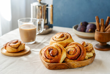 Homemade fresh baked cinnamon rolls in a wooden tray, cinnamon sticks, fig, coffee-pot and cappuccino 