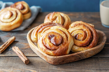 Homemade fresh baked cinnamon rolls in a wooden tray, cinnamon sticks, sugar and coffee with milk over wooden and dark blue background