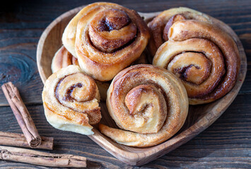 Homemade cinnamon rolls in a wooden tray in a heart shape with cinnamon sticks over wooden background, close up