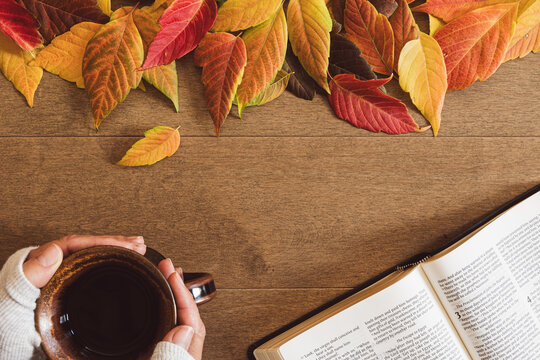Hands holding a cup of coffee by an open bible with a border of autumn leaves on a wood table with copy space