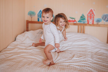 Two little kids, brother and sister, both dressed in white are jumping on a bed covered with a white blanket