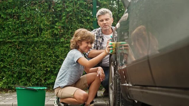 Beautiful family washing their car with sponges in sunny day. Busy father with his hardworking son wiping auto with sponge. People cleaning automobile with foam and water. Helping each other