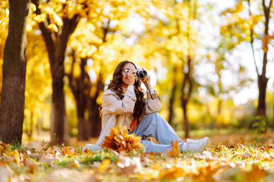 Happy young woman in stylish clothes takes pictures with a retro camera, having fun and relaxing in the autumn sunny park. The concept of relaxation, leisure activities.