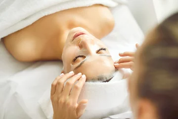 Foto auf Alu-Dibond Spa Professional spa therapist beautician applies white rejuvenating anti age lifting peeling antioxidant detox vitamin sheet mask on face of happy pretty attractive woman for soft smooth good complexion
