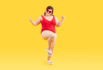 Fototapeta na wymiar Happy plus size woman enjoying summer holiday vacation. Funny fat lady wearing red swimsuit, beach flip flops and sunglasses standing isolated on yellow background, doing YES fist gestures and smiling