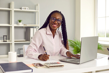 Portrait of friendly dark-skinned female financial advisor working with company accounting data. Woman is sitting at her workplace in front of laptop and calculator and is smiling at camera. - obrazy, fototapety, plakaty