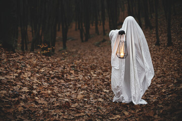 Naklejka premium Spooky ghost holding glowing lantern in moody dark autumn forest. Person dressed in white sheet as ghost with light in evening fall woods. Happy Halloween! Trick or treat