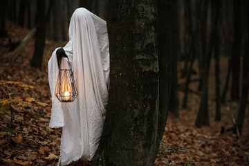 Boo! Happy Halloween! Spooky ghost holding glowing lantern and peeking out of a tree in moody dark...