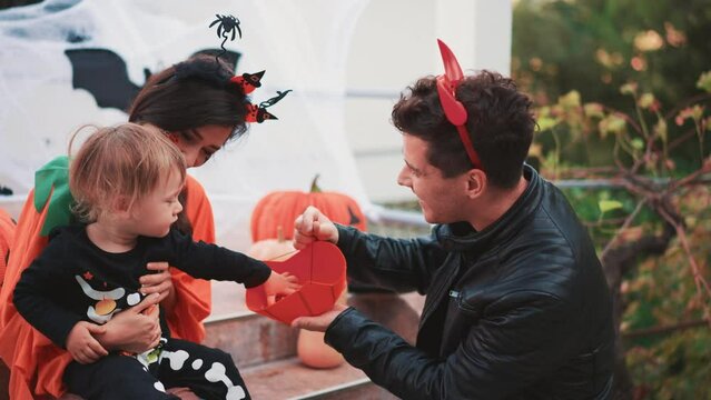Joyous happy mix race Asian family sitting at decorated porch smiling and laughing while playing with carved pumpkins on Halloween. Share collected sweets. Dad mother and cute baby celebrate together