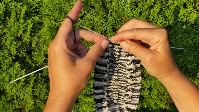 Hands of woman holding wires and hand knitting from black and white melange cotton yarn on sunny weather against of thuja green - timelapse. Topics: hobbies, art, handicrafts, free time, relaxation