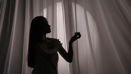 Medium side view shot of a young woman's silhouette wrapped in a towel holding a jar of body, face cream and applying the substance on her skin in a muffled light.