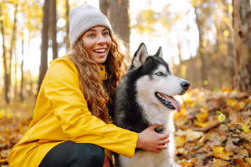 Beautiful young woman in a yellow coat walks in an autumn park with her pet husky. A pet owner spends time with her dog. Concept of fun, entertainment.