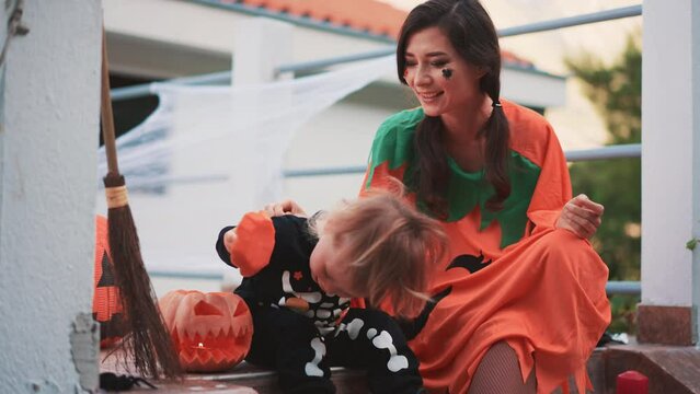 Joyous happy mix race Asian family portrait sitting at decorated porch smiling and laughing while playing with pumpkins on Halloween. Mother and cute baby celebrate autumn tradition holiday together