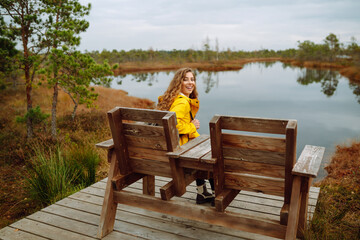 Smiling traveler in a bright coat with a backpack walks along a wooden path in the swamps. Travel concept, exploring. Active lifestyle.