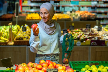 Portrait of a female buyer in a supermarket, a Muslim woman in a hijab chooses an apple fruit, happy with a large assortment of products and smiles, woman shopping in a large grocery store.