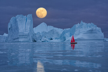 Red sailboat sailing among floating icebergs in front of the full moon rising at Artic Ocean in...